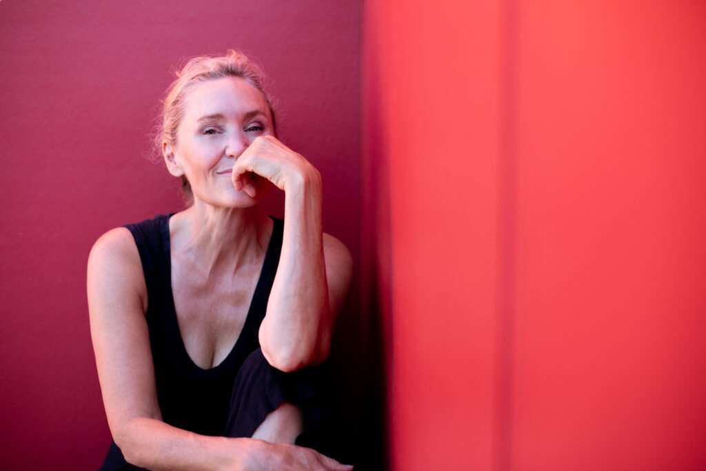 Woman sitting against a bright red wall with her hand raised.