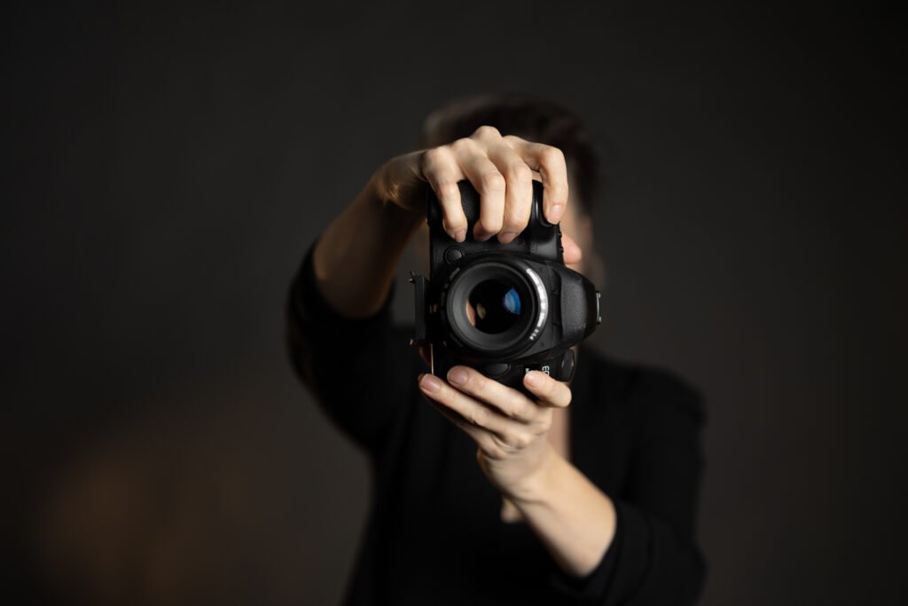 Photographer holding a camera directly toward the viewer against a dark background