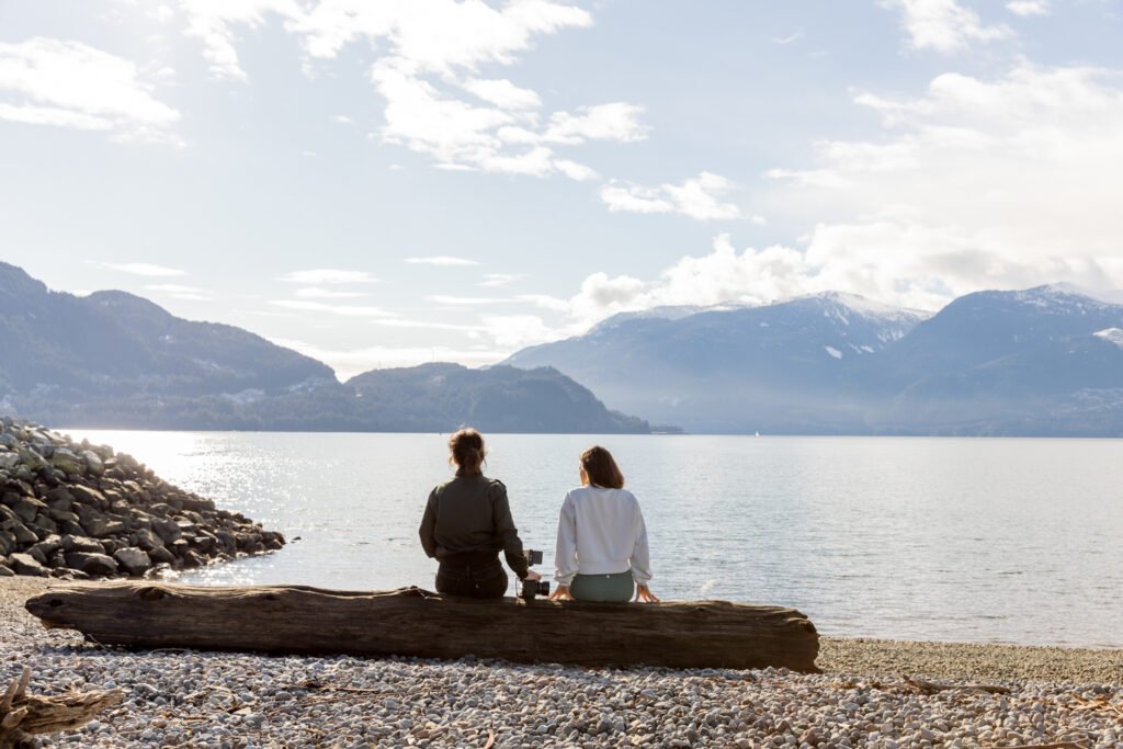 Two women sitting on a log beside a mountain lake.