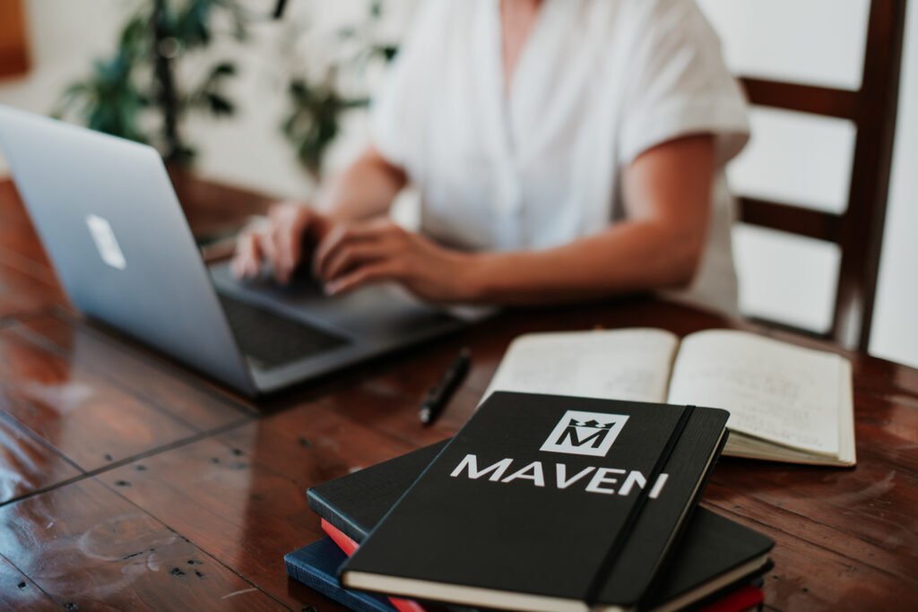 Person working on a laptop beside notebooks and branded Mavene books.