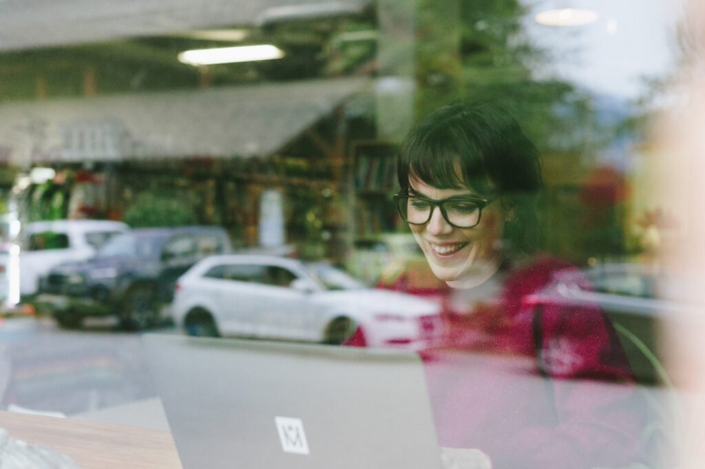 Woman smiling while working on a laptop near a window.